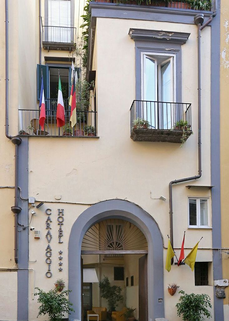 Entrada de un hotel con un letrero que dice 'HOTEL CARAVAGGIO', balcones con plantas y banderas.