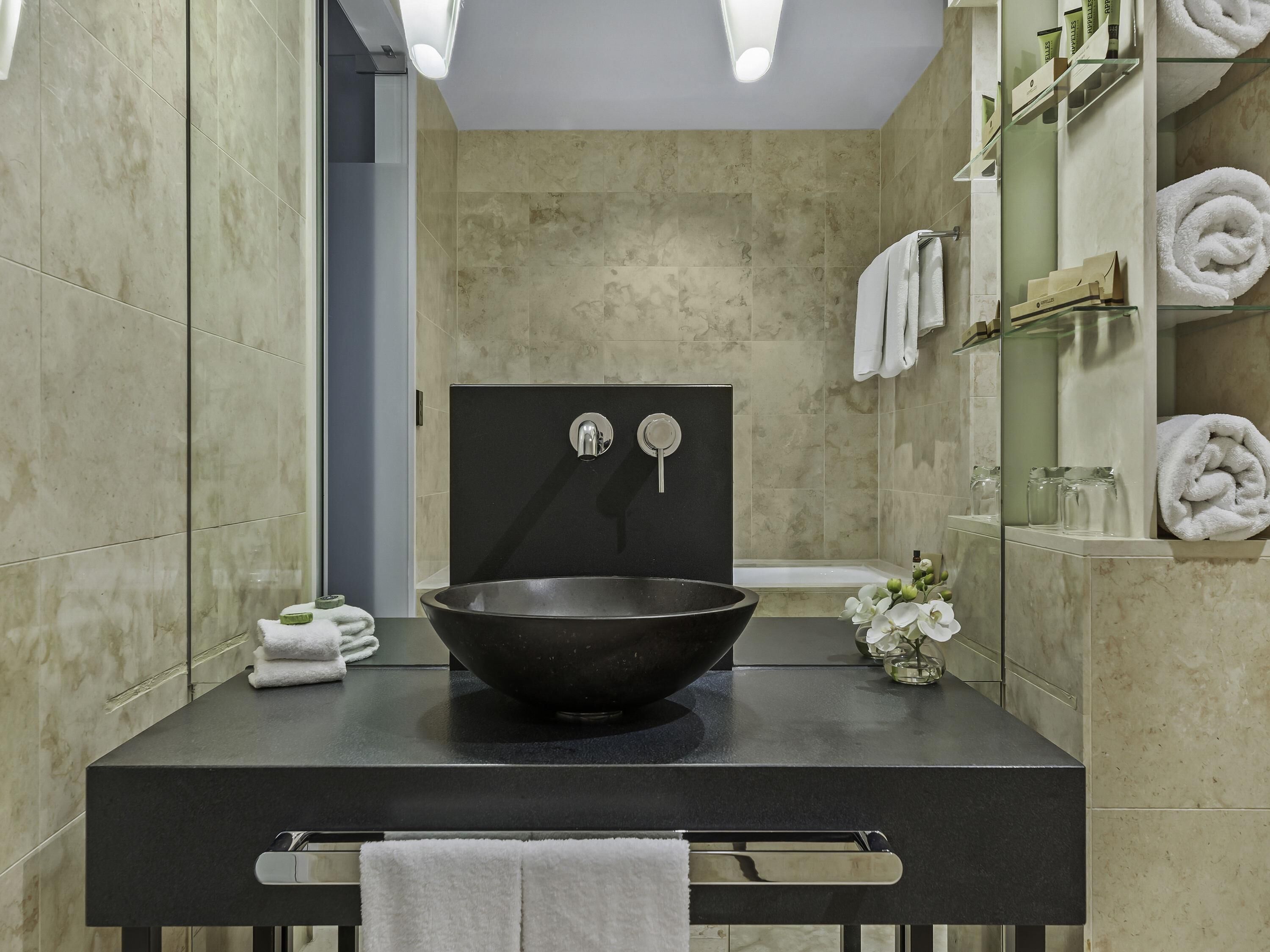 Un baño moderno con un lavamanos negro, un espejo y una repisa de vidrio con artículos de aseo.