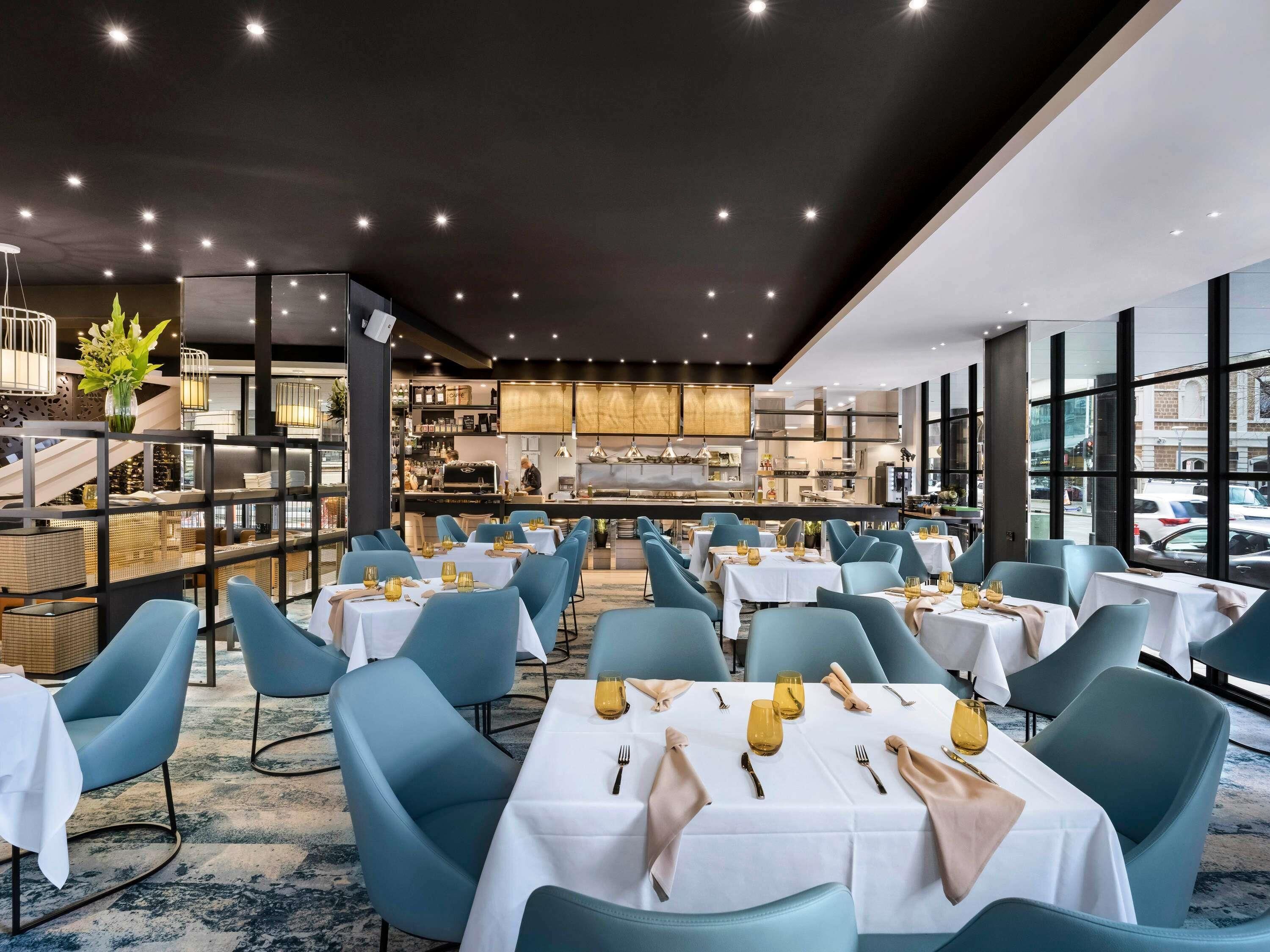 A restaurant with tables set for dining, a kitchen in the background, and large windows.