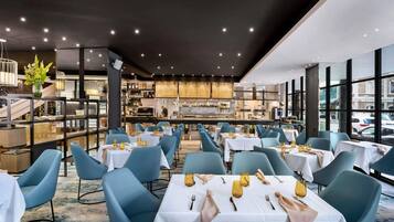 A restaurant with tables set for dining, a kitchen in the background, and large windows.