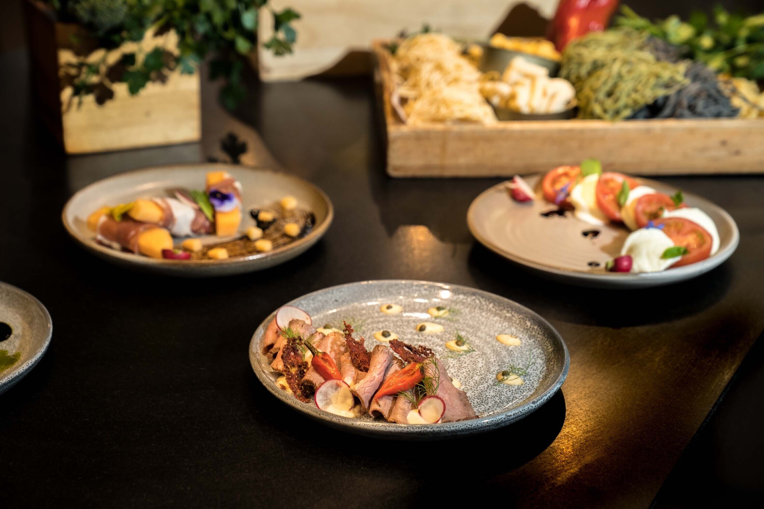 Une table à manger avec trois assiettes de nourriture, une boîte en bois contenant du pain et une plante en arrière-plan.