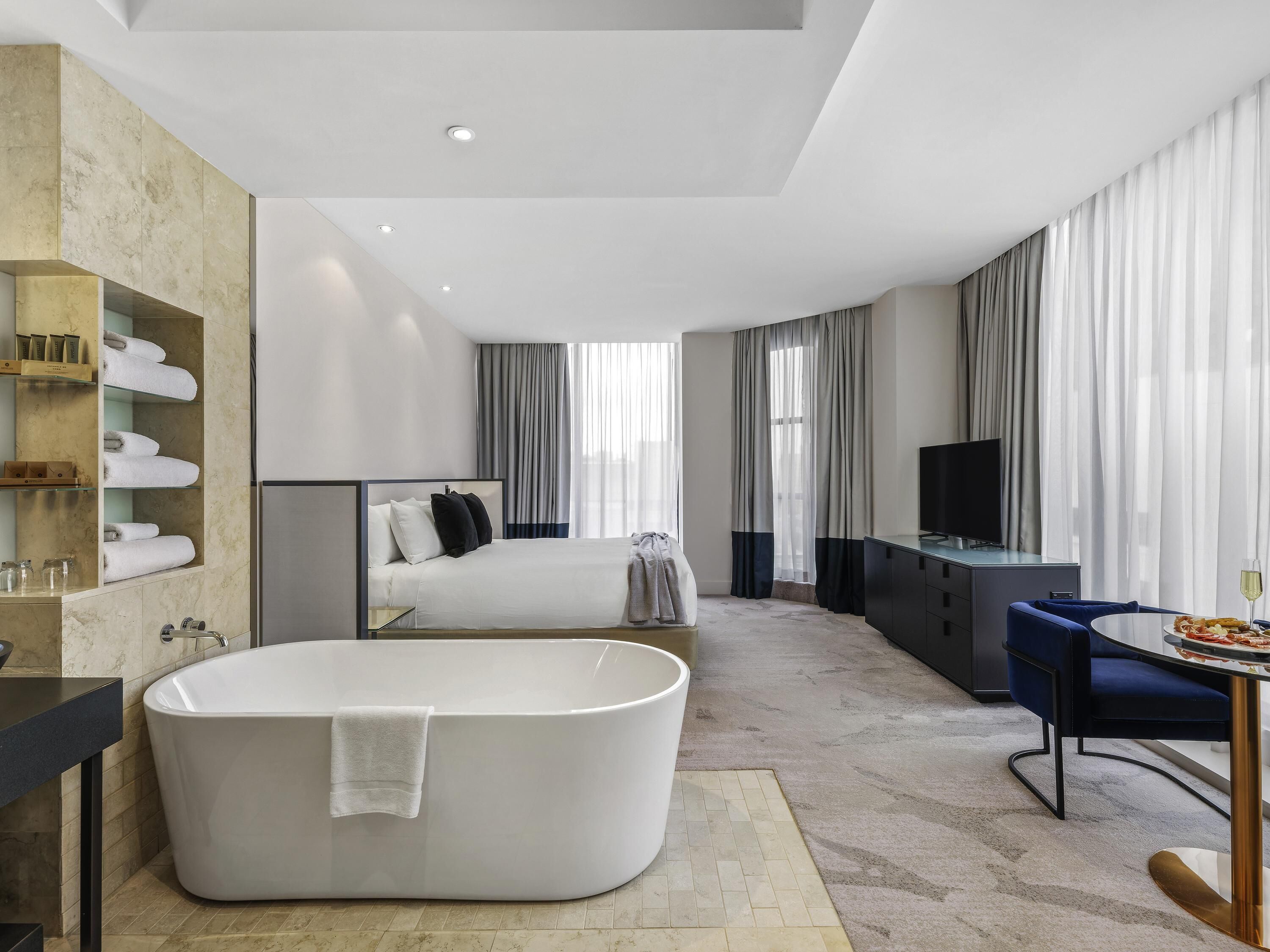 A modern hotel room with a large bathtub, a bed, a desk with a chair, and a television.