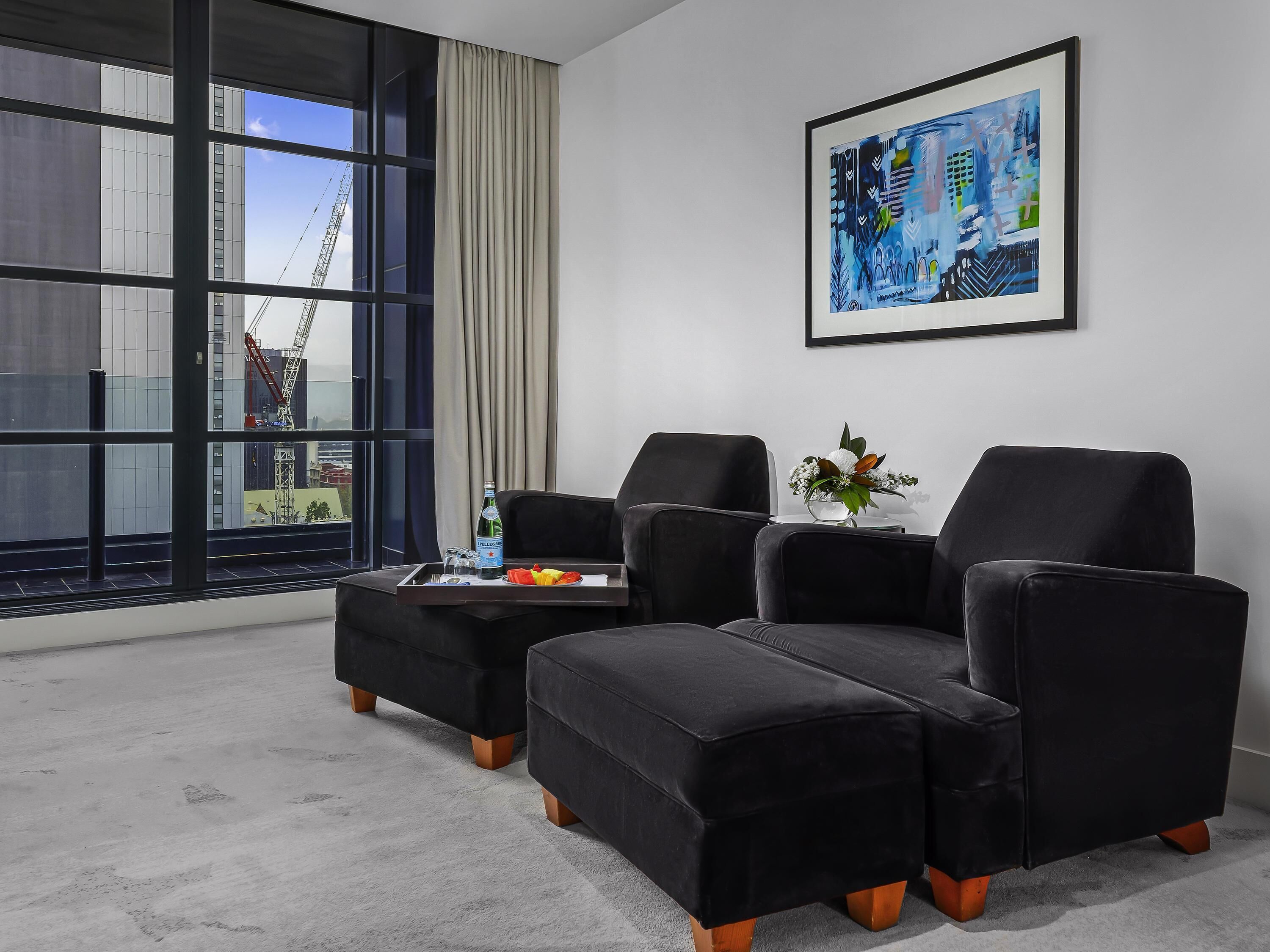 A modern hotel room with a large window, black upholstered chairs, a small table with a tray, and a framed abstract painting on the wall.