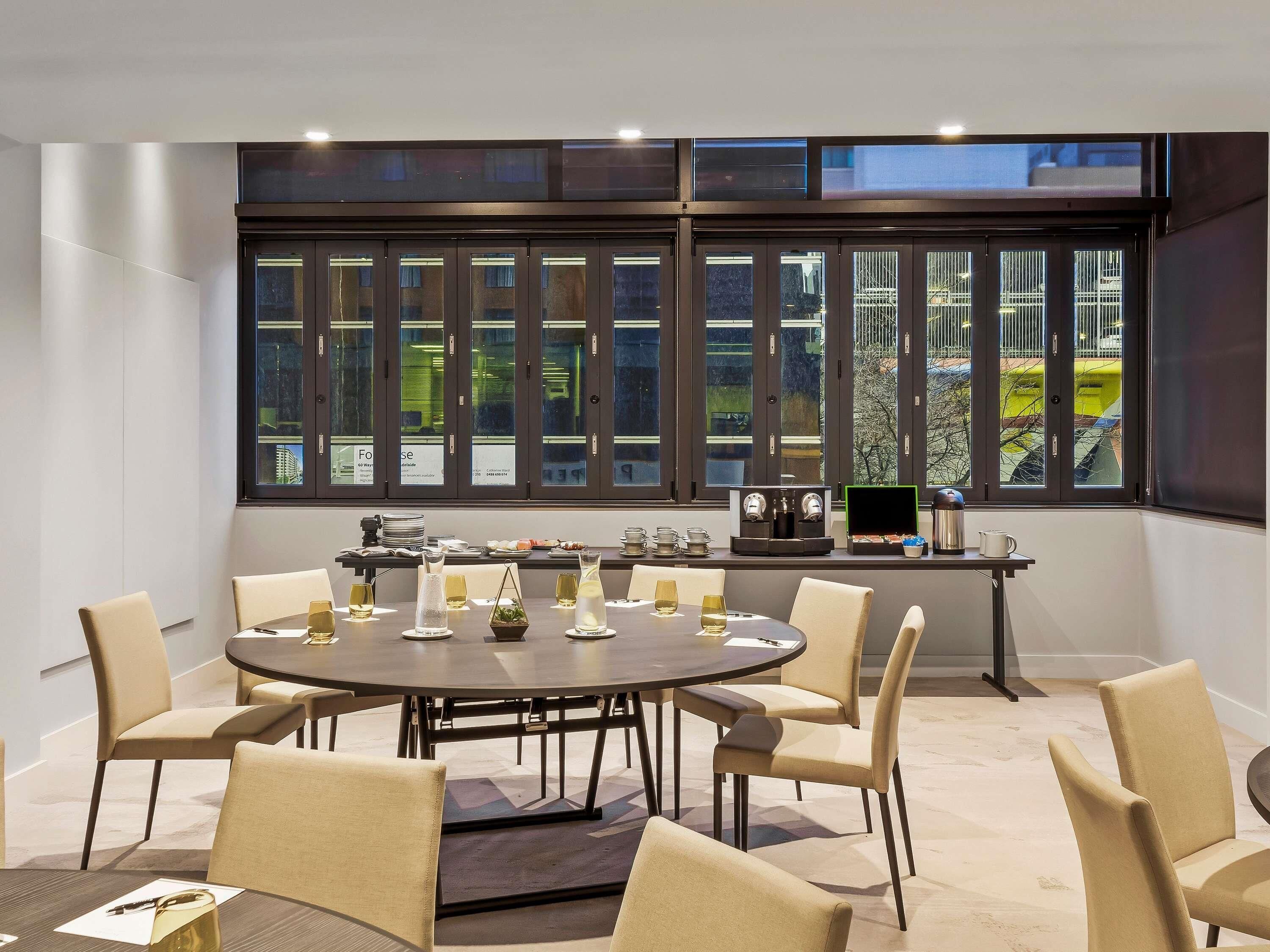 A conference room with a round table, chairs, a coffee machine, and a large window with a view of greenery.