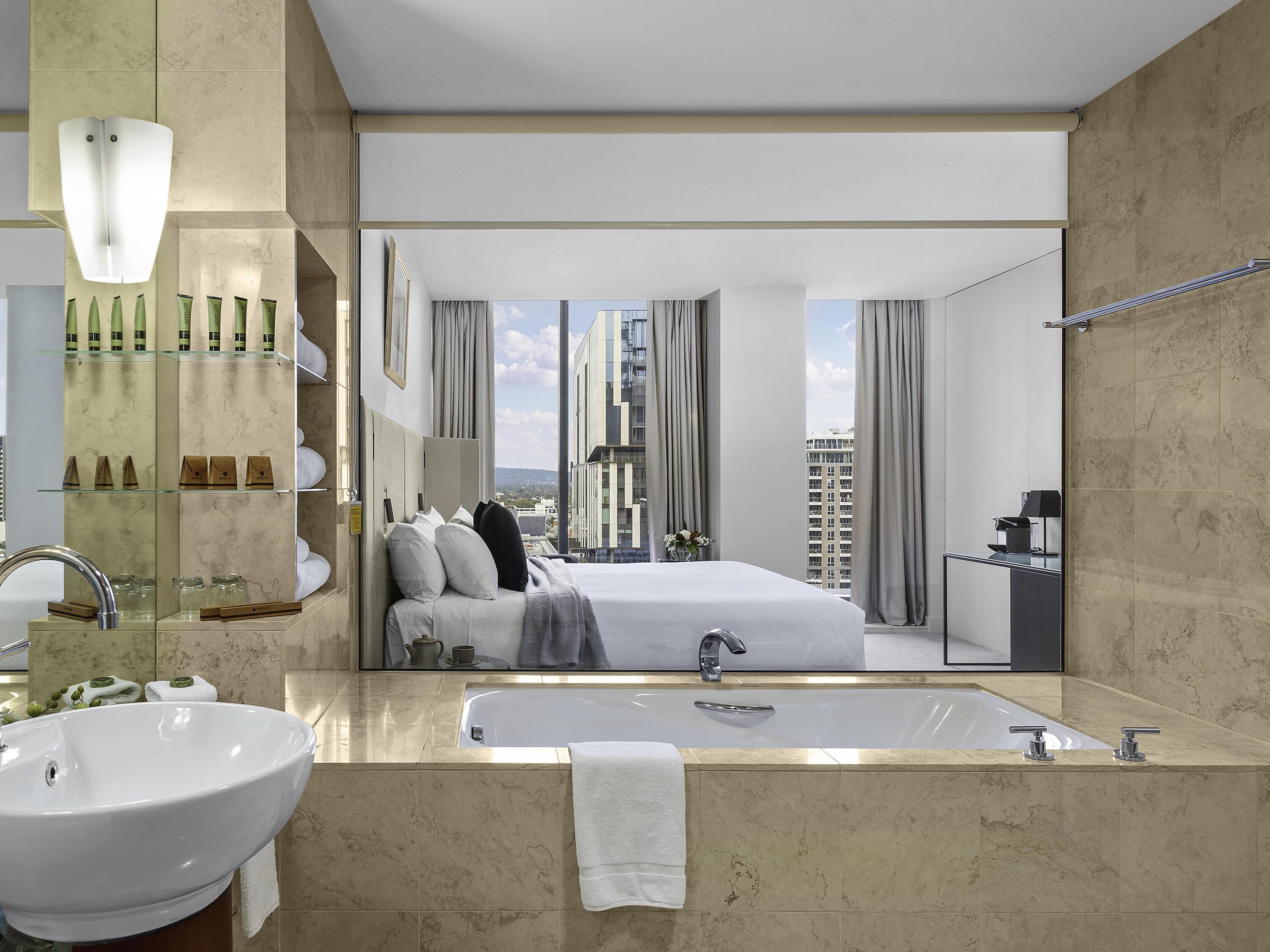A modern hotel room with a large bed, a bathtub, a vanity with a sink, and a view of the cityscape.