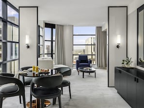 A modern hotel room with a dining area, a seating area with a blue armchair, and a view of the cityscape through large windows.