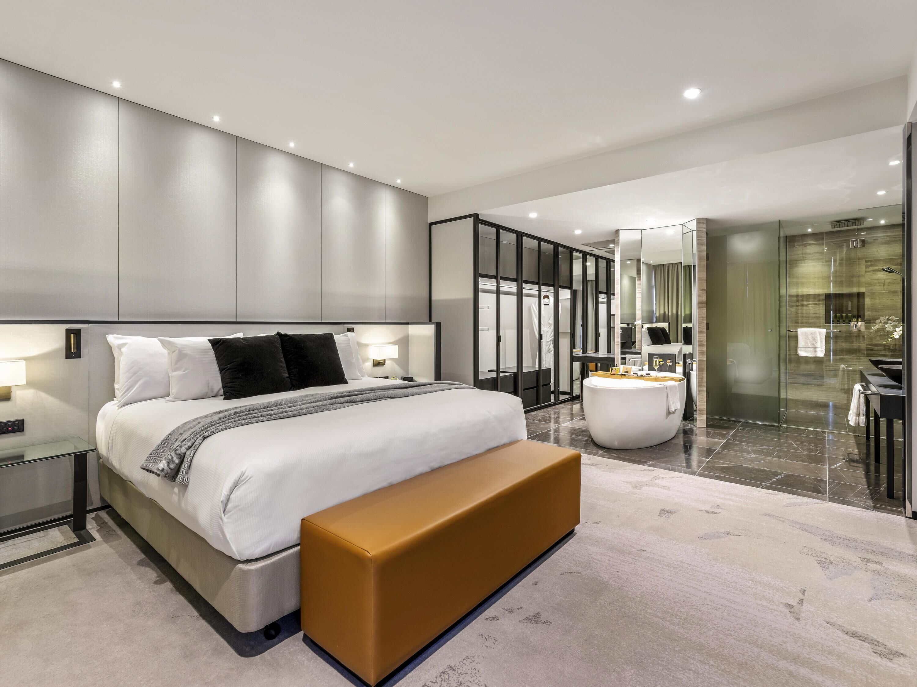 Una habitación de hotel moderna con una cama grande, mesitas de noche, un banco, un vestidor, un baño con ducha de vidrio y tocador.