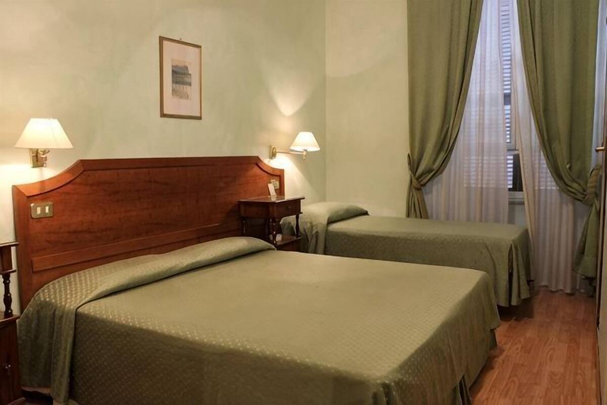 Camera d'albergo con due letti, una testiera in legno, lampade sui comodini, un quadro appeso al muro e una finestra con tende.
