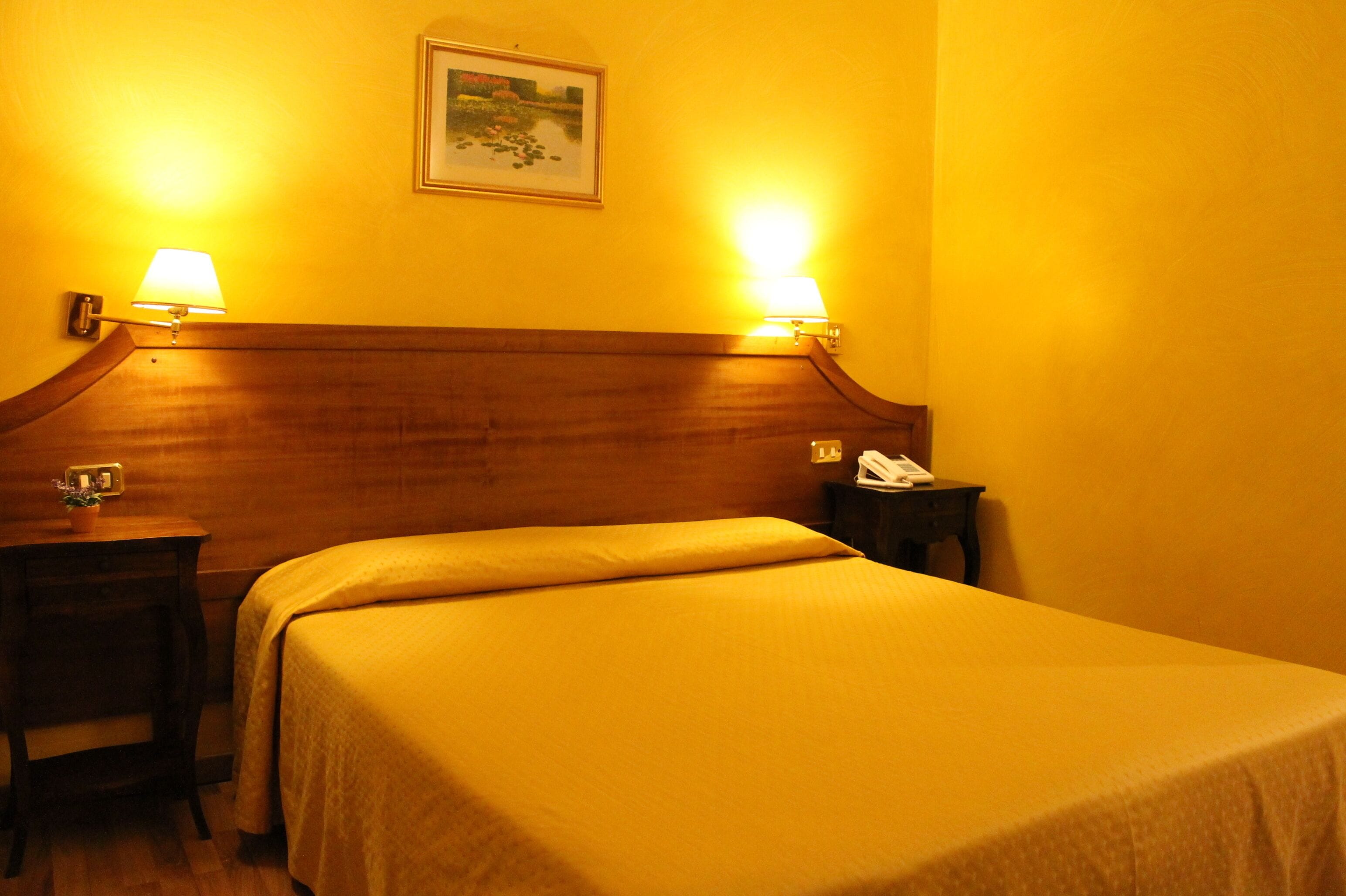 Habitación de hotel con cabecera de madera, dos lámparas de noche, una mesita de noche con un teléfono y un cuadro enmarcado en la pared.
