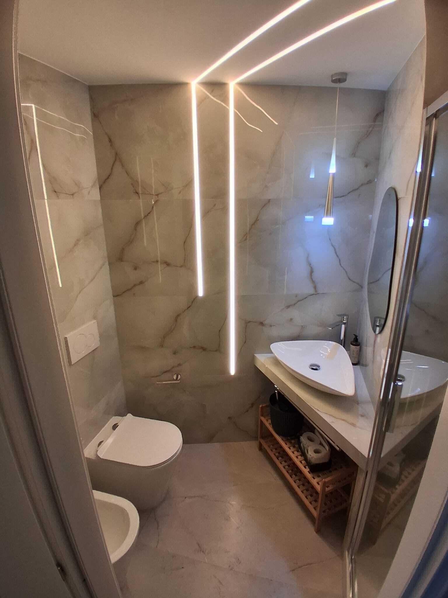 A modern bathroom with a marble wall, a white sink, a toilet, and a glass shower enclosure.