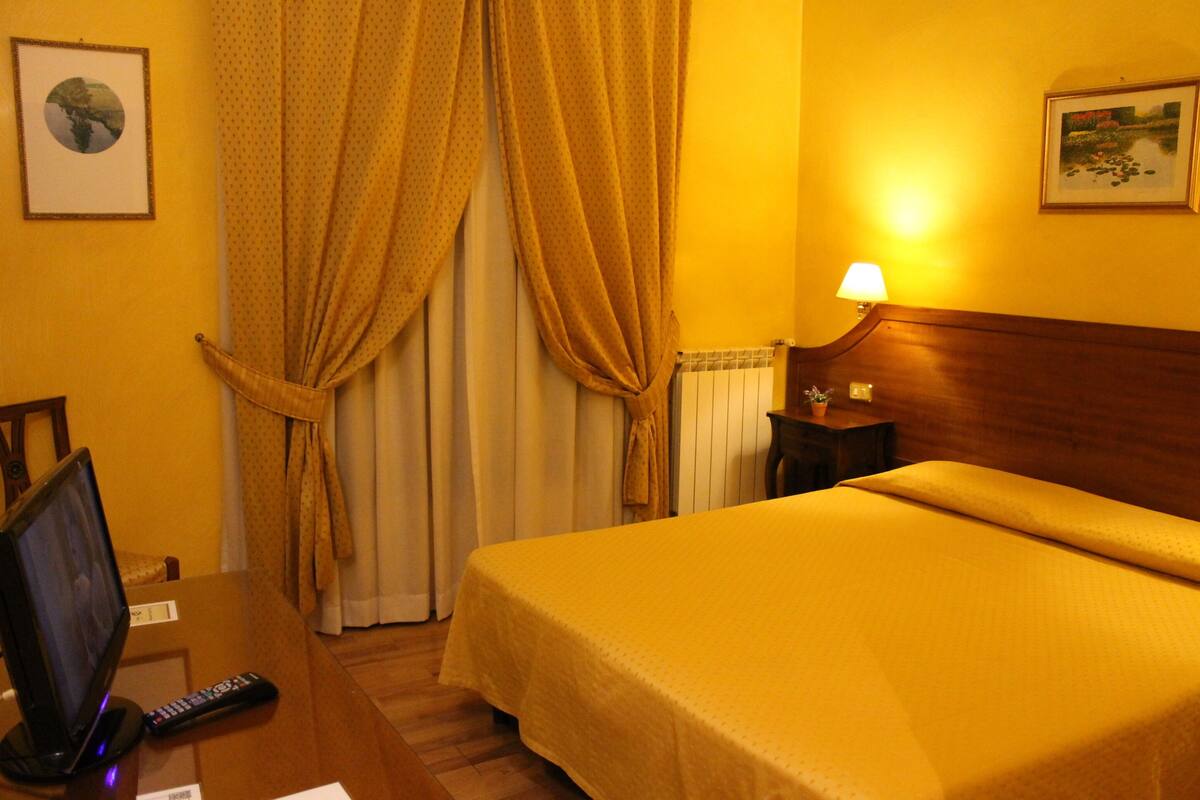 Una camera d'albergo con un letto, una scrivania con un monitor computer e un telecomando, una lampada e tende.