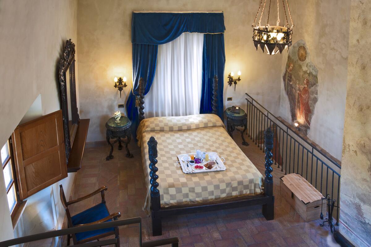A bedroom with a checkered bedspread, a wooden chair, a bedside table with a tray of breakfast items, and a large painting on the wall.