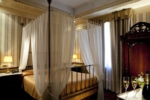 Quarto de hotel com cama de dossel, mesa de cabeceira com abajur e bandeja com duas taças de vinho e uma garrafa.