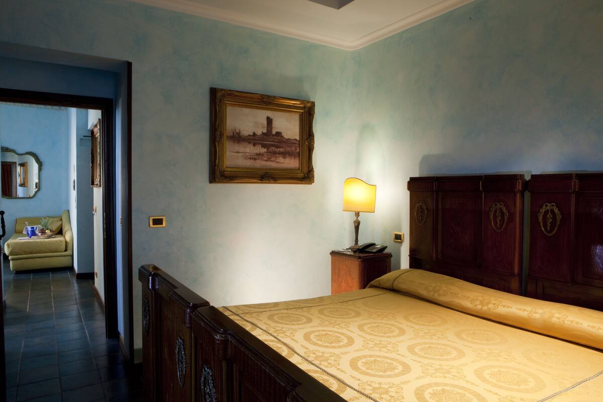 A bedroom with a bed, wooden headboard, a nightstand with a lamp, a framed picture on the wall, and a view of a hallway with a sofa.