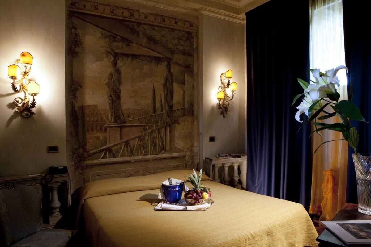 A hotel room with a bed, a wall mural, a tray with fruits and a mug, and a vase with flowers.