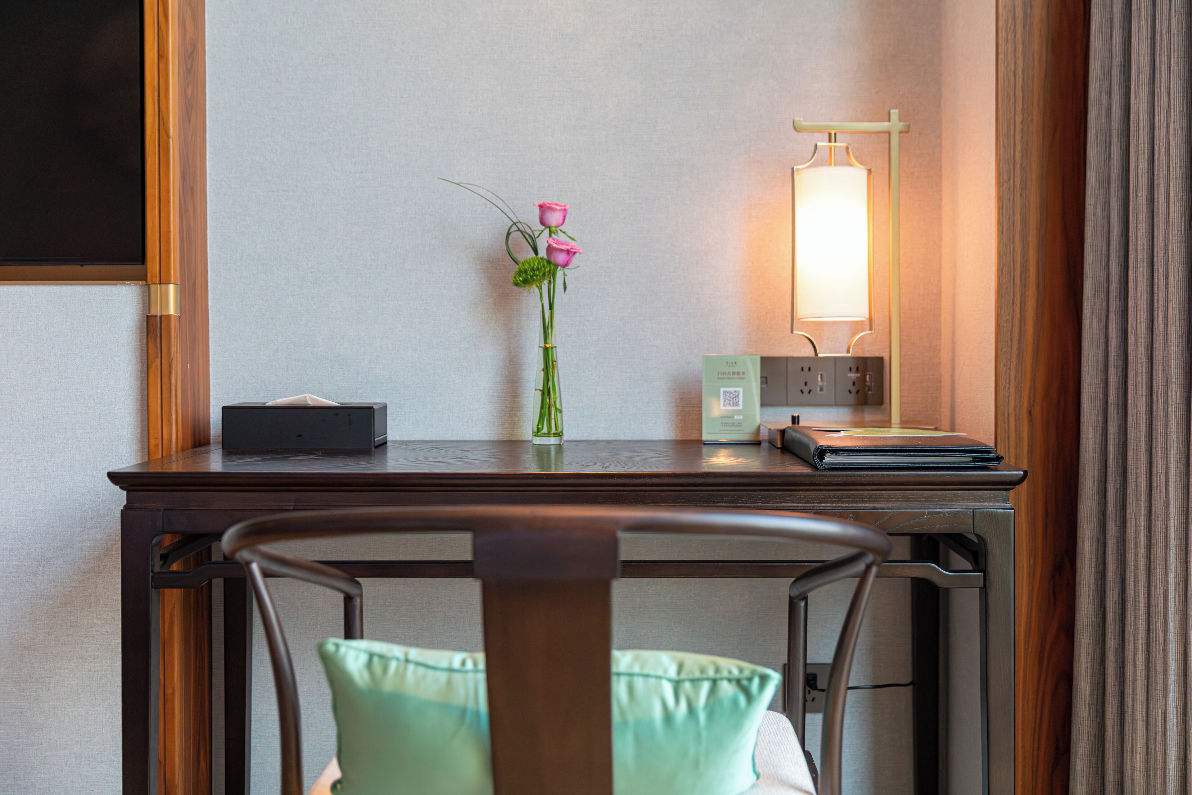 Et hotelværelse med et træskrivebord, en vase med lyserøde blomster, en sengetavlelampe og en stol med grøn pude.