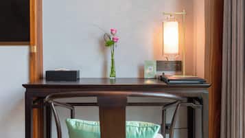 A hotel room with a wooden desk, a vase with pink flowers, a bedside lamp, and a chair with a green cushion.
