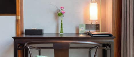 A hotel room with a wooden desk, a vase with pink flowers, a bedside lamp, and a chair with a green cushion.