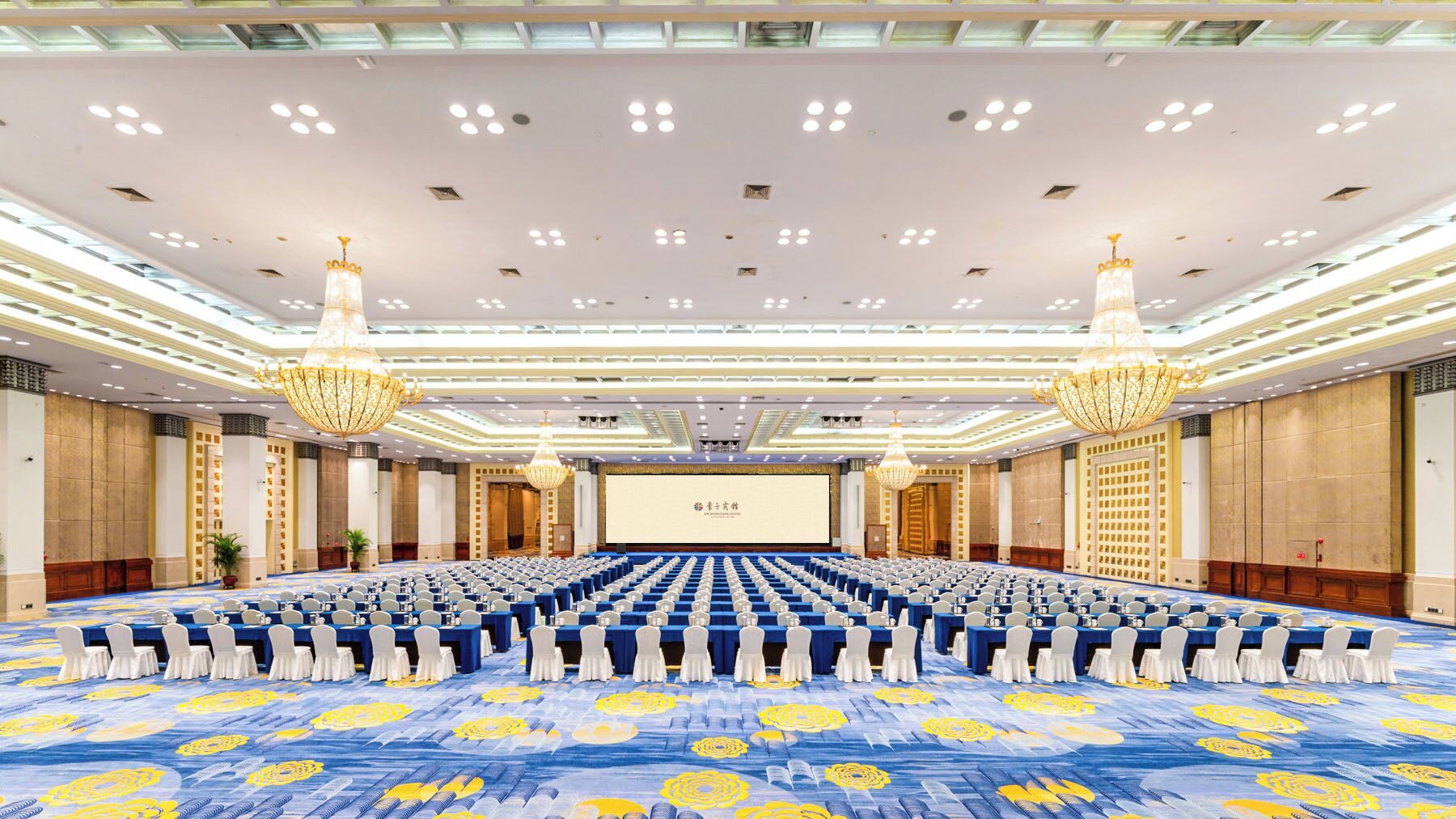 A large conference room with rows of tables and chairs, a stage with a screen, and chandeliers.