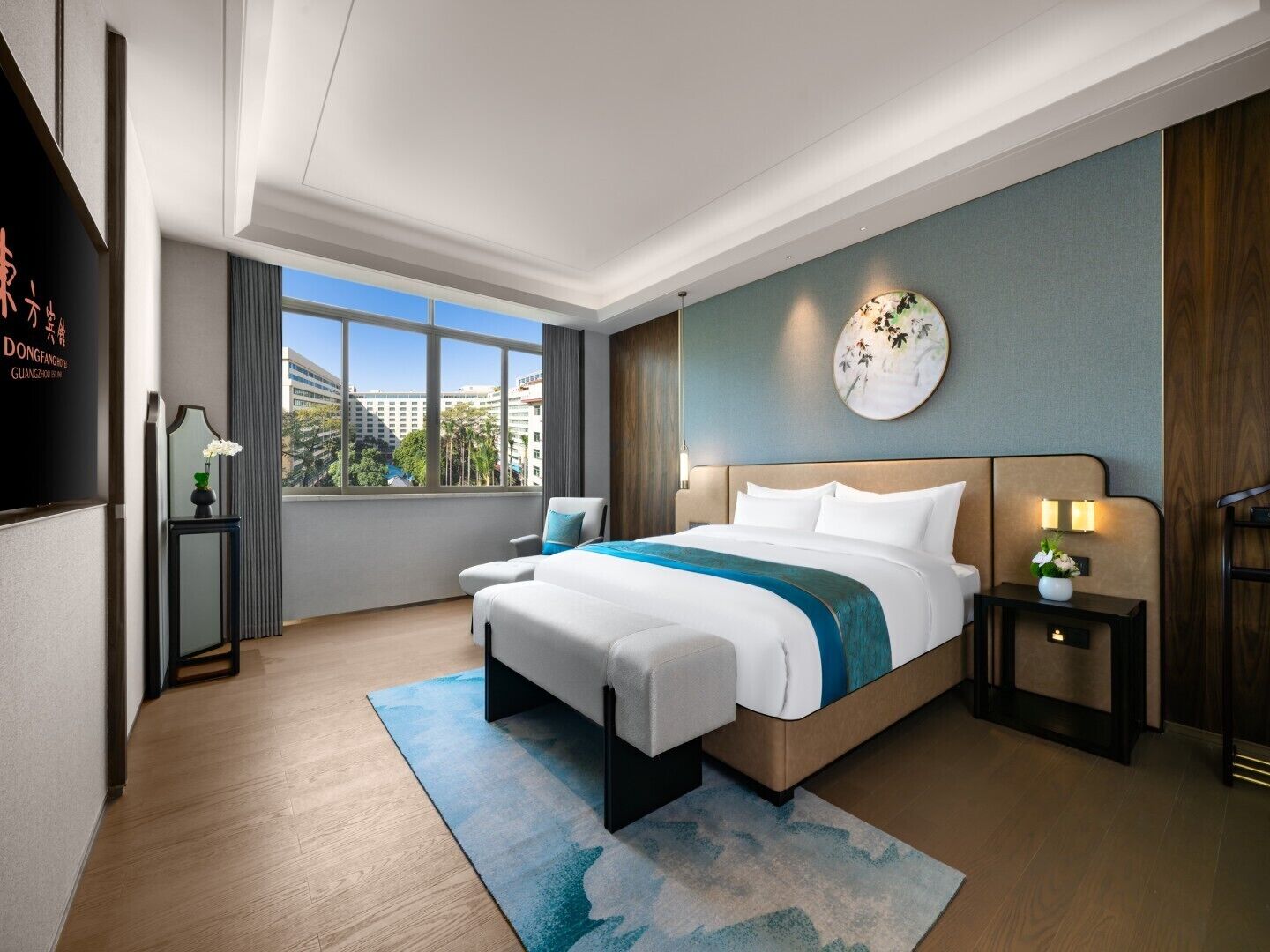 Una moderna camera d'hotel con un letto grande, un comodino con una lampada, vista sulla città attraverso ampie finestre e un elemento decorativo a parete.