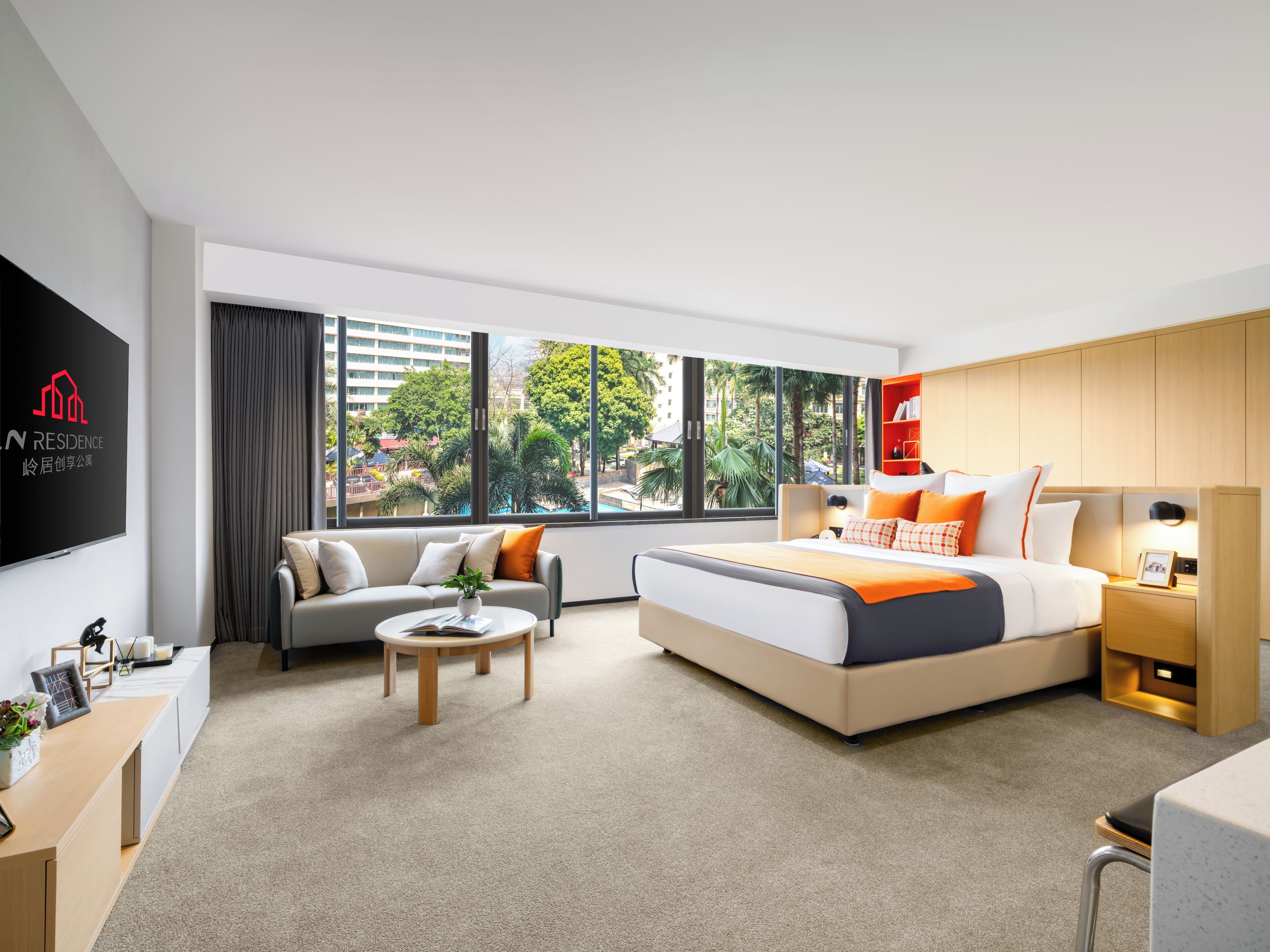 A modern hotel room with a large bed, a sofa, a coffee table, and a TV. The room has large windows with a view of buildings and trees.