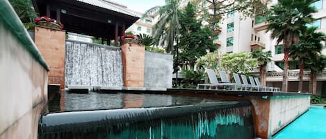 A pool with a waterfall feature, lounge chairs, and a building with a tiled roof in the background.