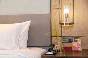A hotel room with a bed, a bedside table with a phone, a vase with a plant, and a wall-mounted light fixture.