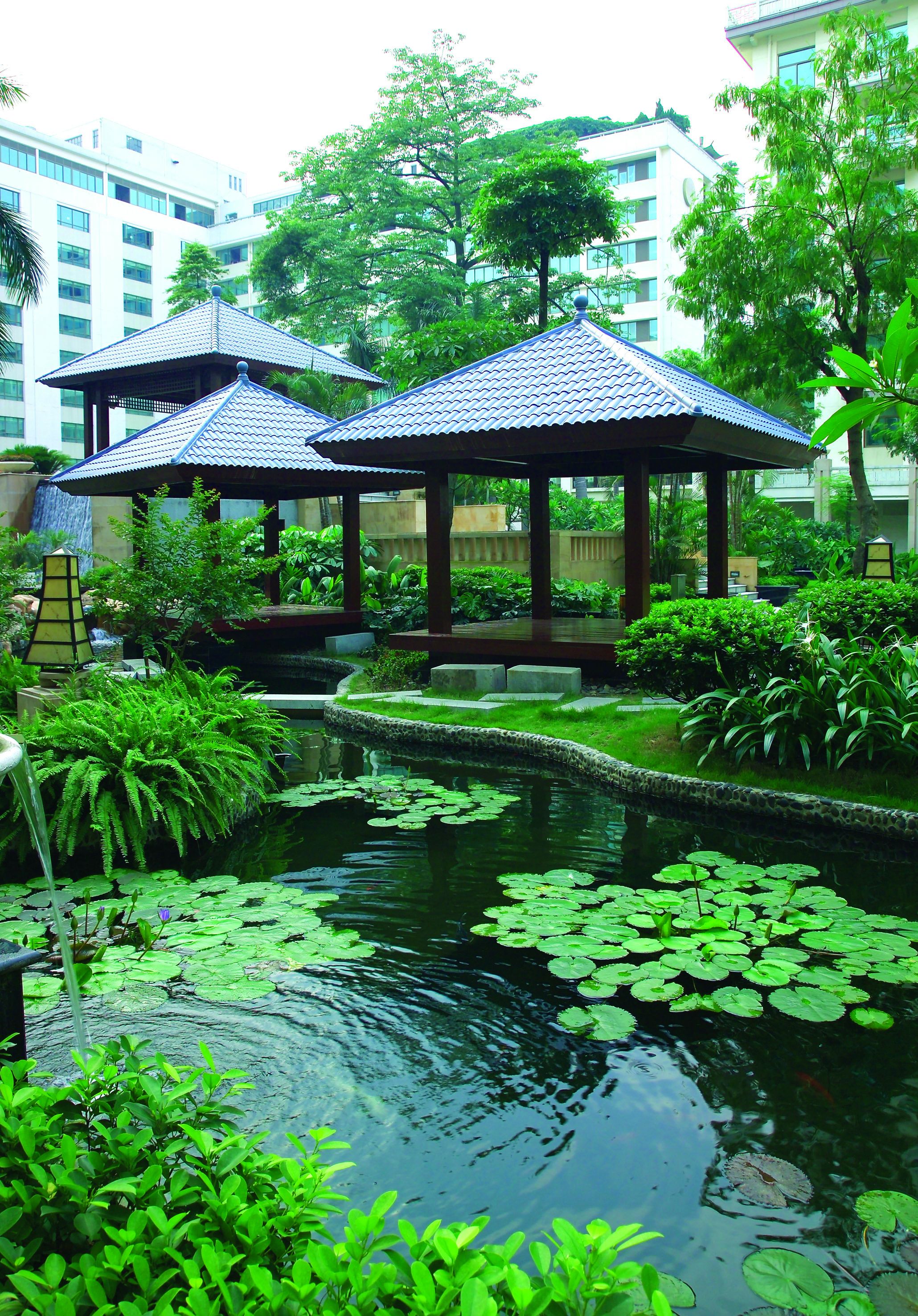 A tranquil garden with a pond, surrounded by lush greenery and traditional-style pavilions.