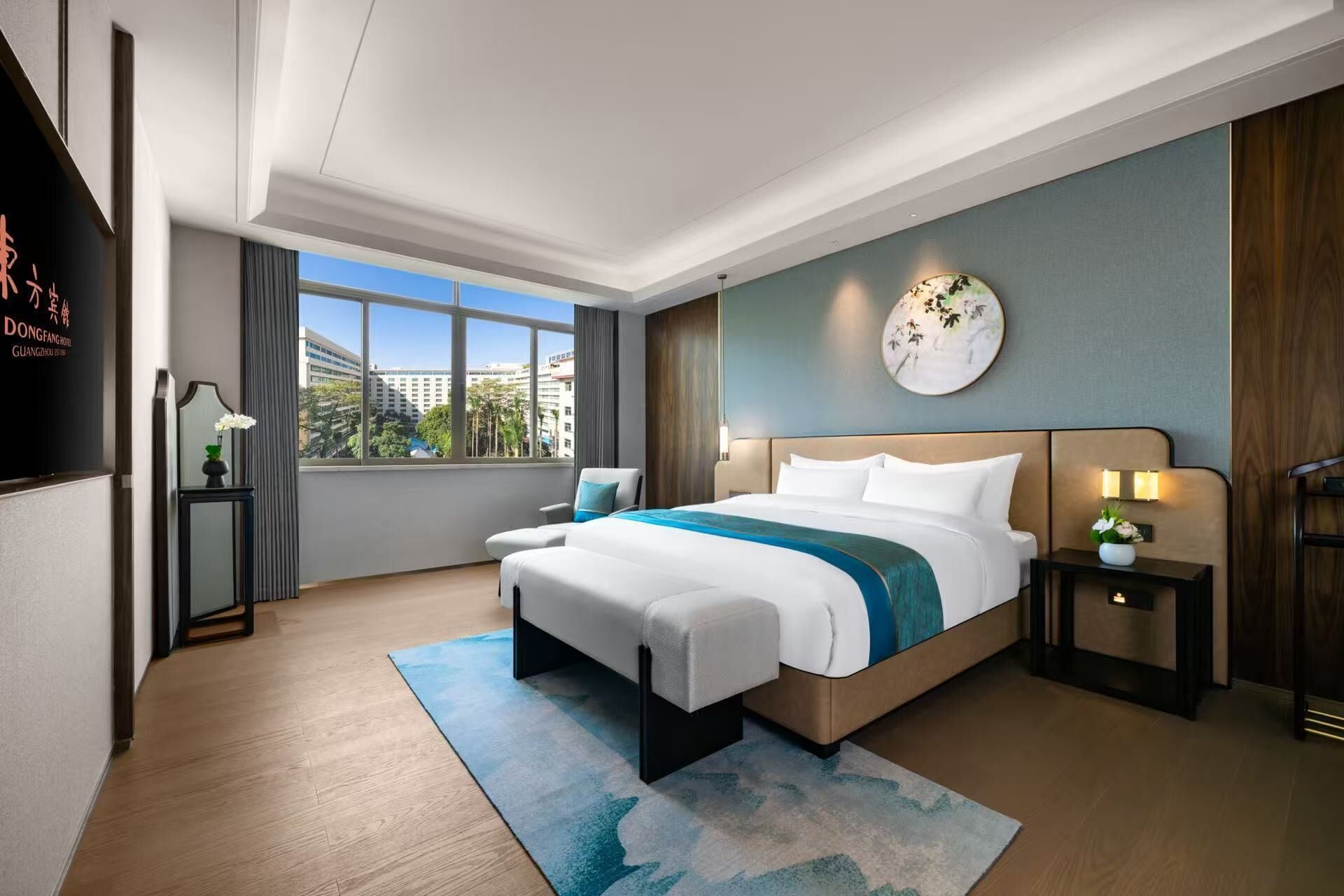 Habitación de hotel con una cama grande, una mesita de noche, un reloj de pared y una ventana con vistas a la ciudad.