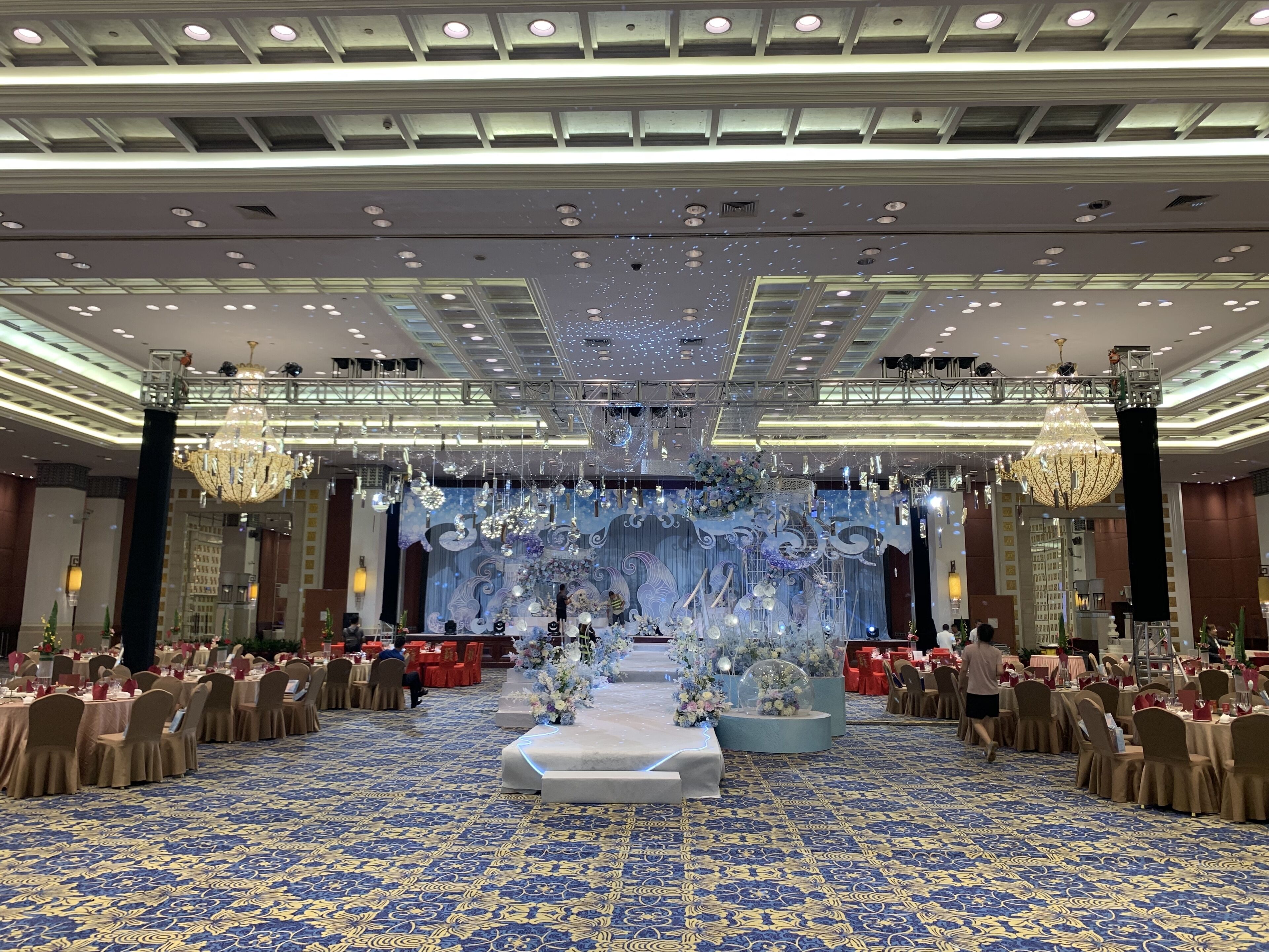 Un salón de banquetes con alfombra azul estampada, un escenario con adornos florales y candelabros.