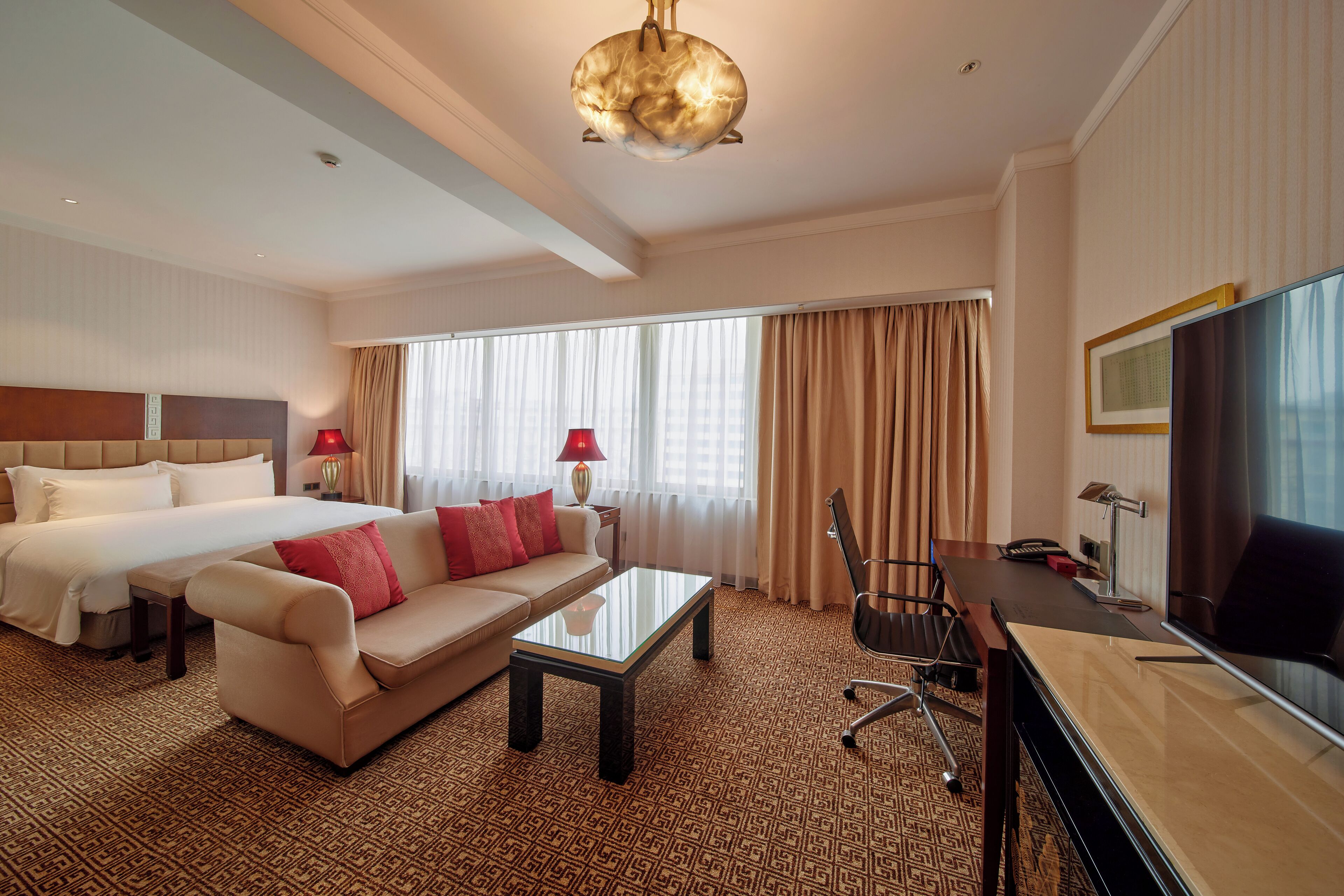 A hotel room with a bed, sofa, coffee table, desk, TV, and large windows with curtains.