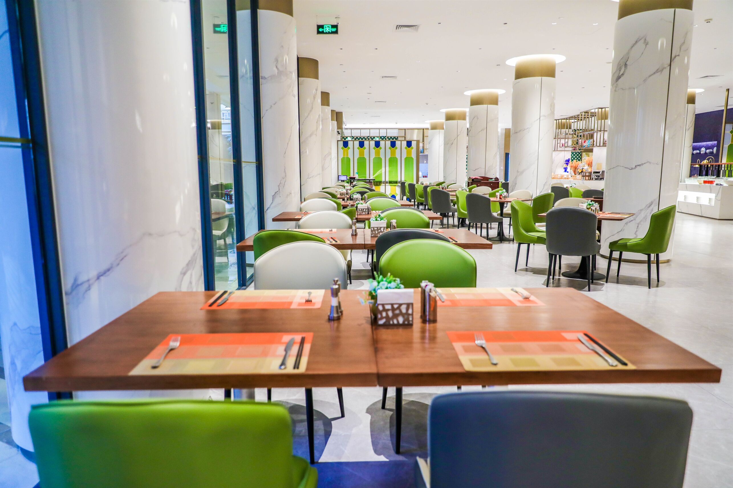 A modern restaurant with marble columns, green and grey chairs, and neatly arranged tables with cutlery.