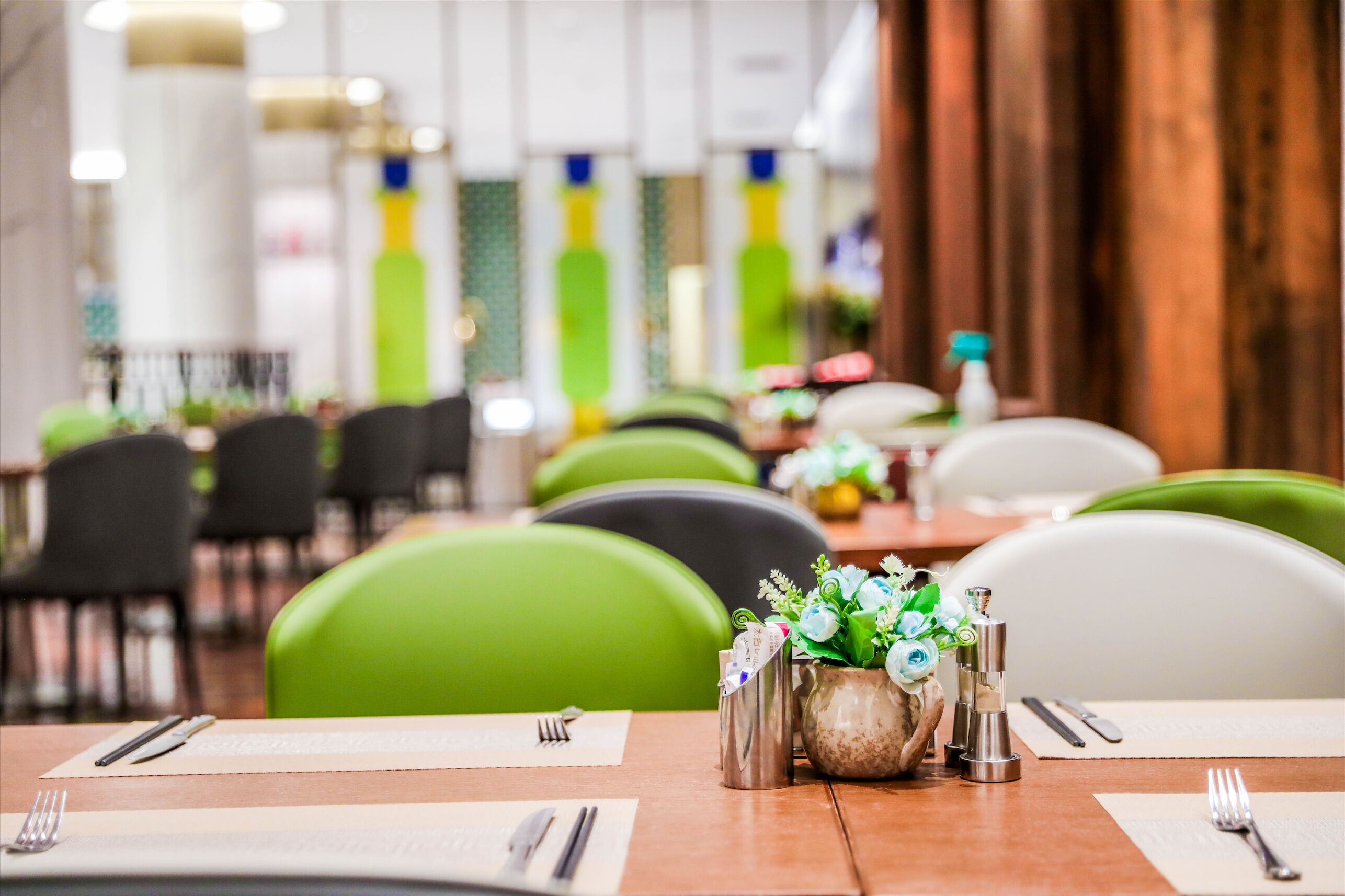 A restaurant with green chairs, neatly set tables with cutlery, and a vase with flowers.