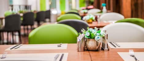 Un restaurante con sillas verdes, mesas ordenadas con cubiertos y un jarrón con flores.