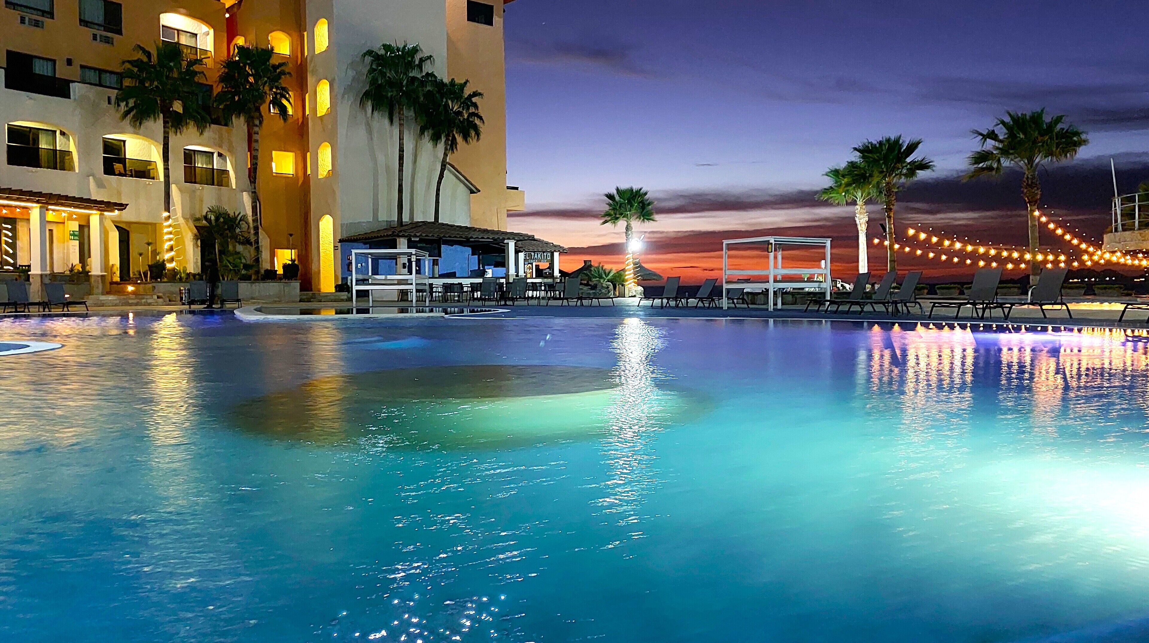 A swimming pool with lounge chairs and palm trees, a building with illuminated windows, and a sunset in the background.