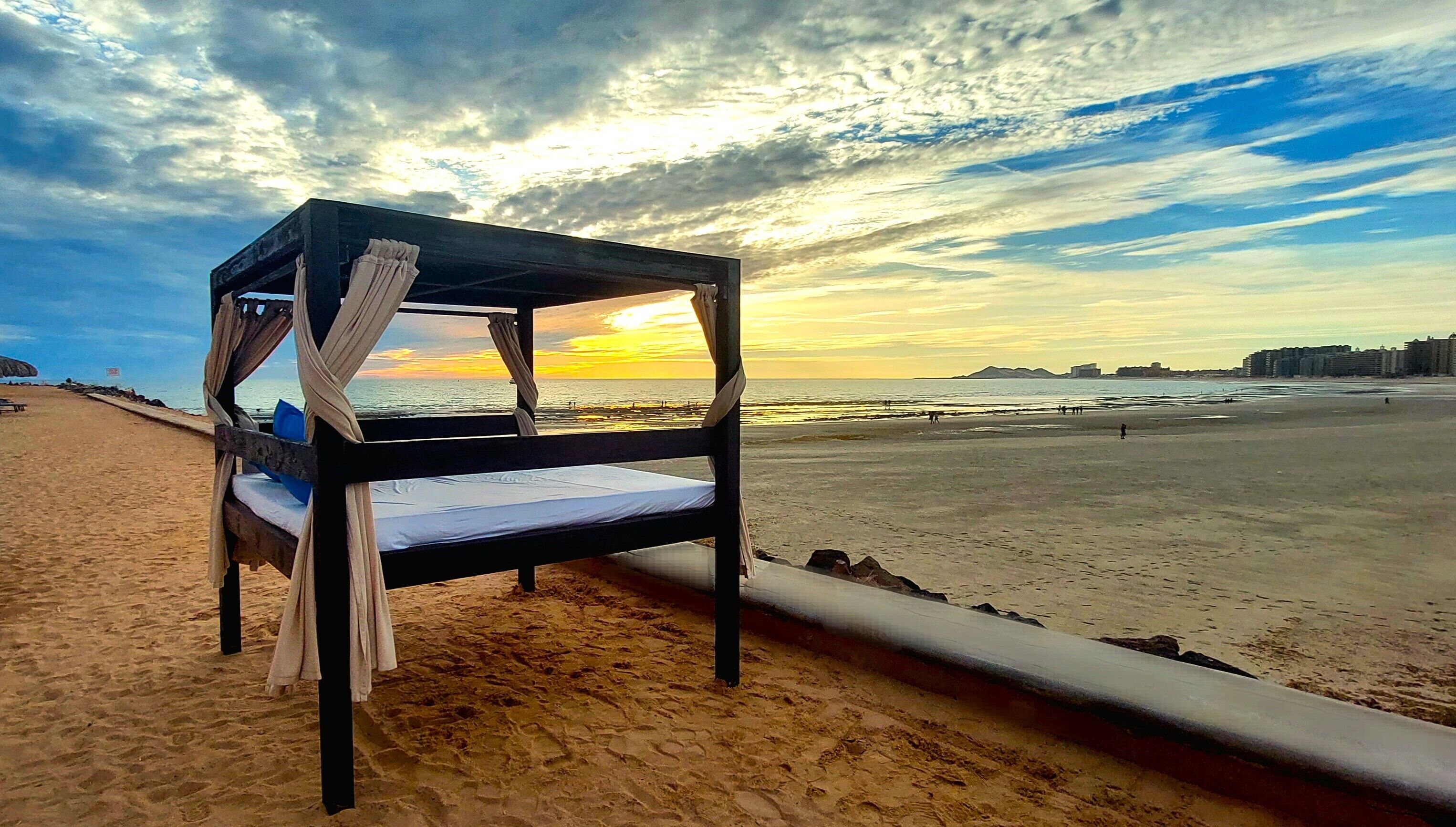A beach view with a wooden bed structure, a sandy beach, and a sunset in the background.