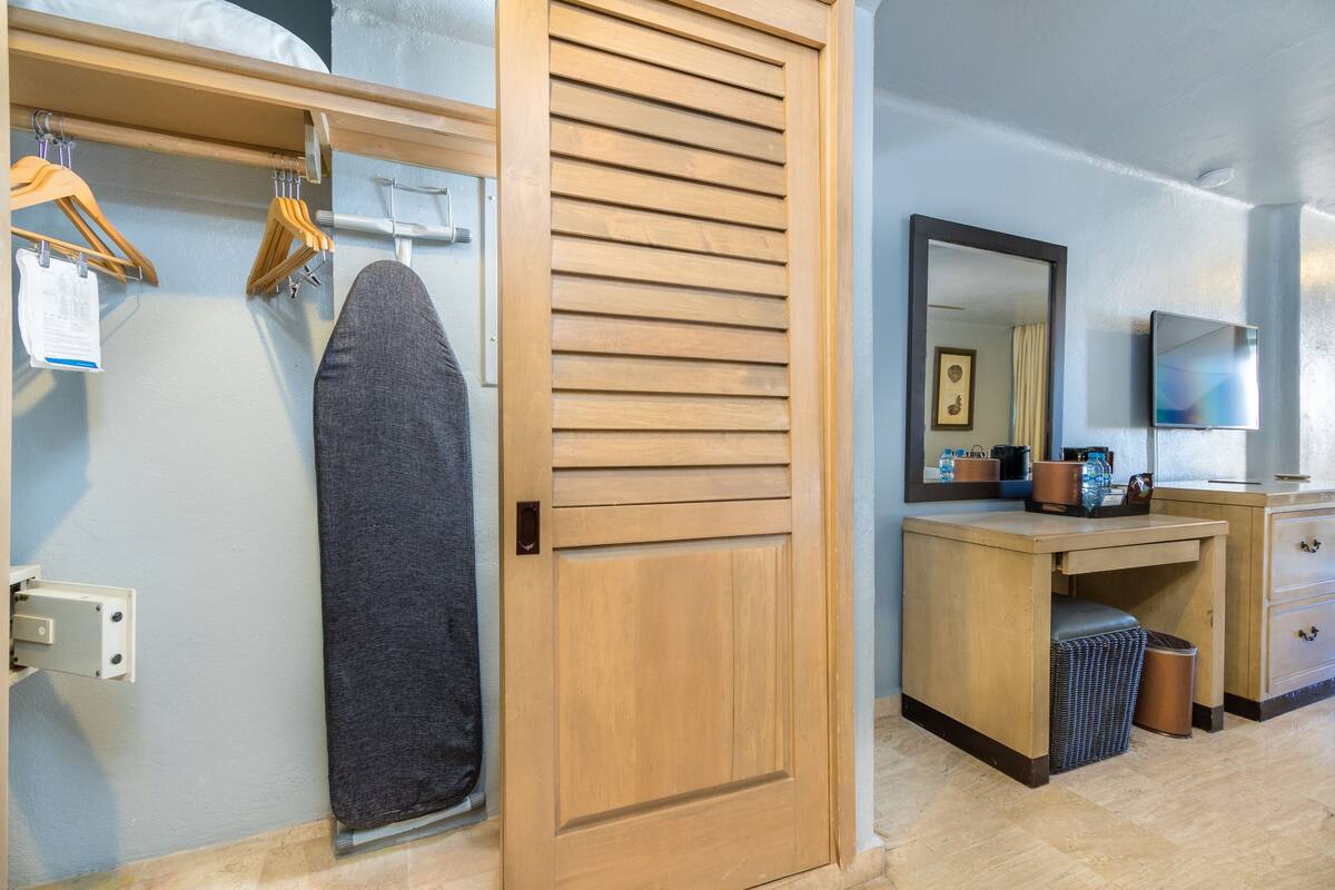 A hotel room with a wooden wardrobe, a mirror, a television, and a small desk with a trash bin.