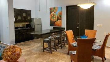 Cocina compacta con armarios oscuros, microondas y refrigerador. Hay una zona de comedor con sillas de mimbre y una mesa de vidrio, y un cuadro de vegetales en la pared.