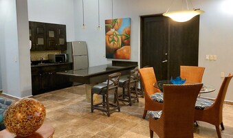 Cocina compacta con armarios oscuros, microondas y refrigerador. Hay una zona de comedor con sillas de mimbre y una mesa de vidrio, y un cuadro de vegetales en la pared.