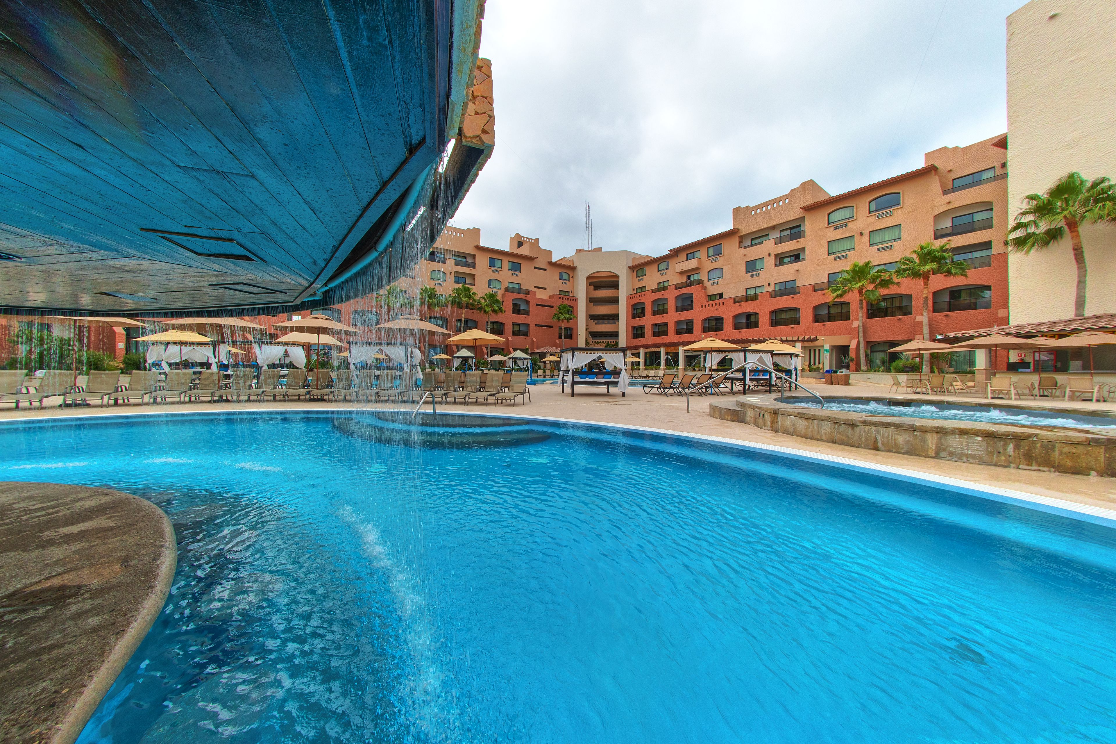 A large swimming pool with multiple lounge areas and umbrellas, surrounded by multi-story buildings with balconies and palm trees.
