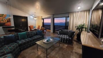 A modern living room with a sofa, armchair, and coffee table. There is a TV, a painting, and a view of the ocean.