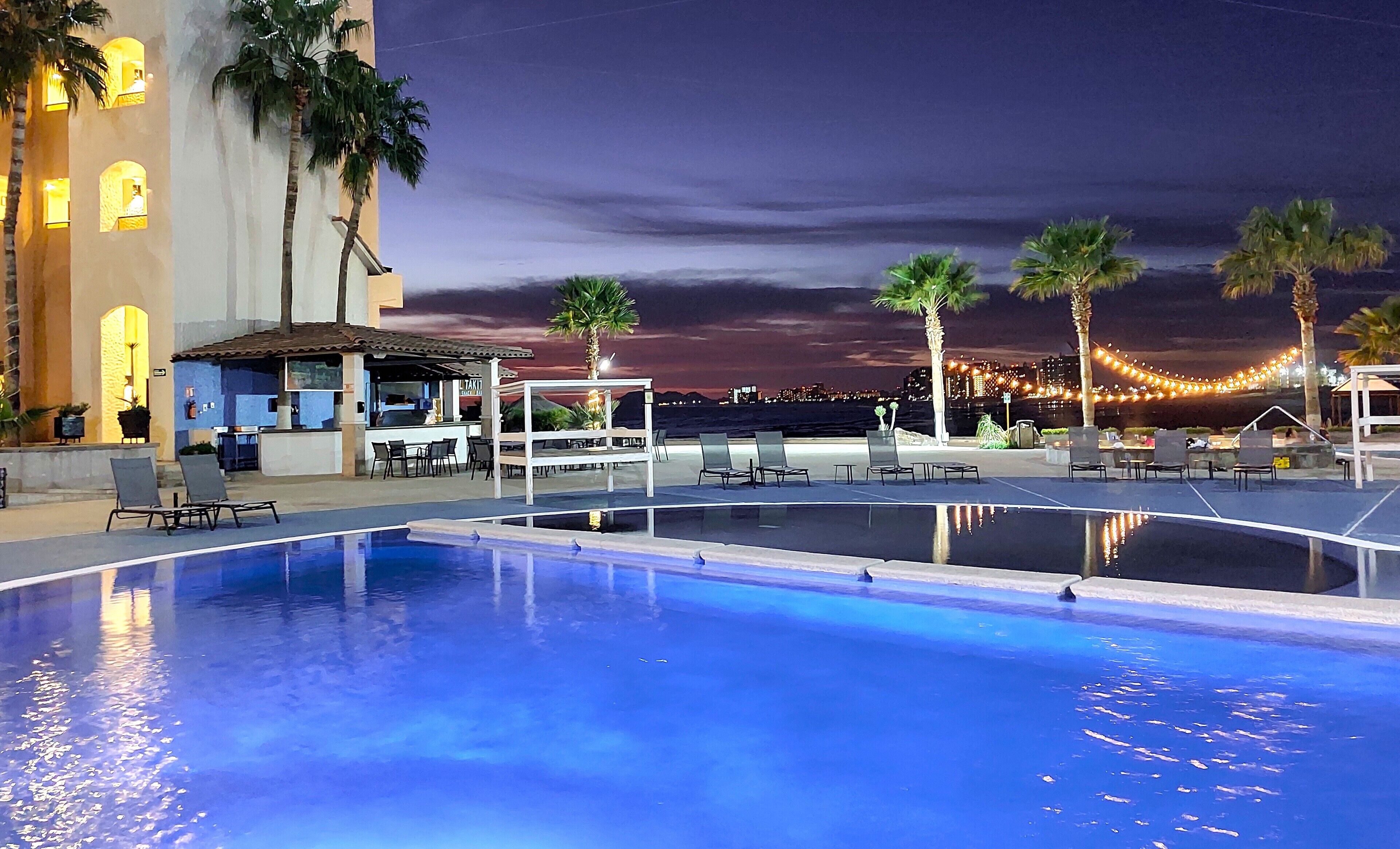 Un espace piscine doté de chaises longues et d’un bar, offrant une vue sur le paysage urbain au crépuscule.