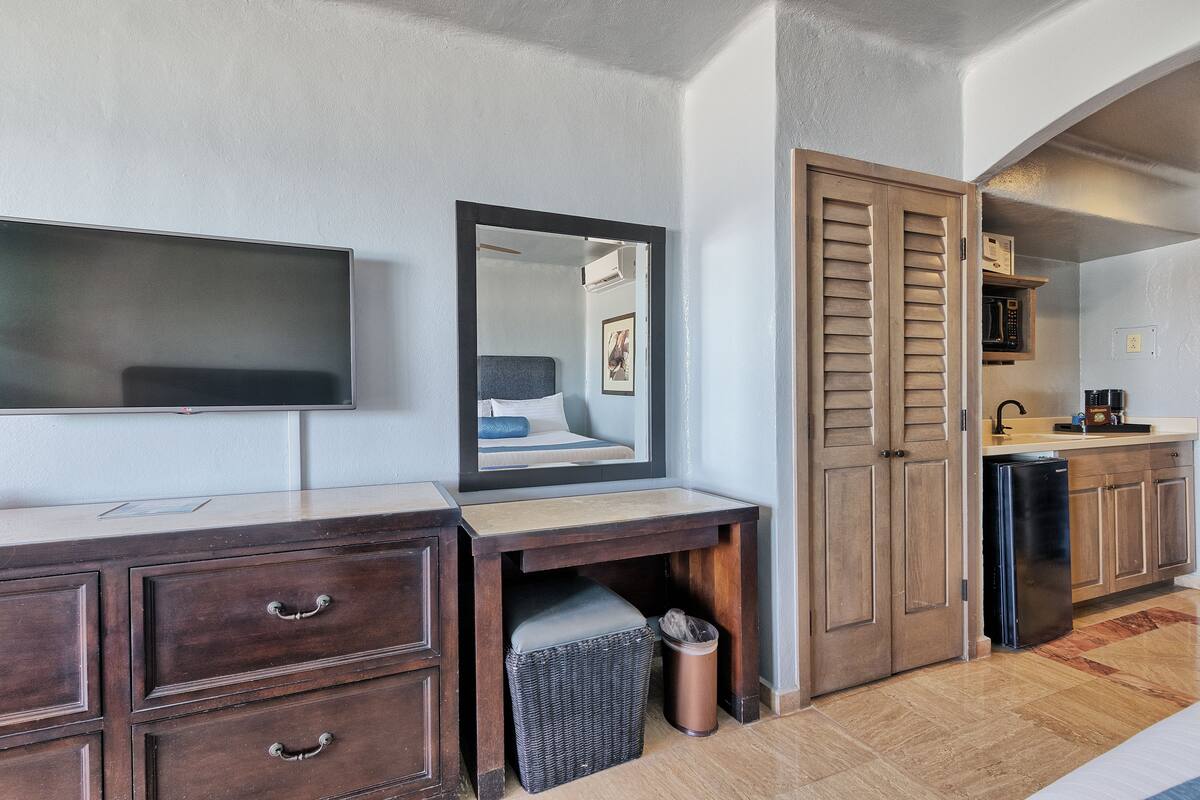 A modern hotel room with a wooden desk, a flat-screen TV, a mirror, a small trash bin, and a compact kitchenette.