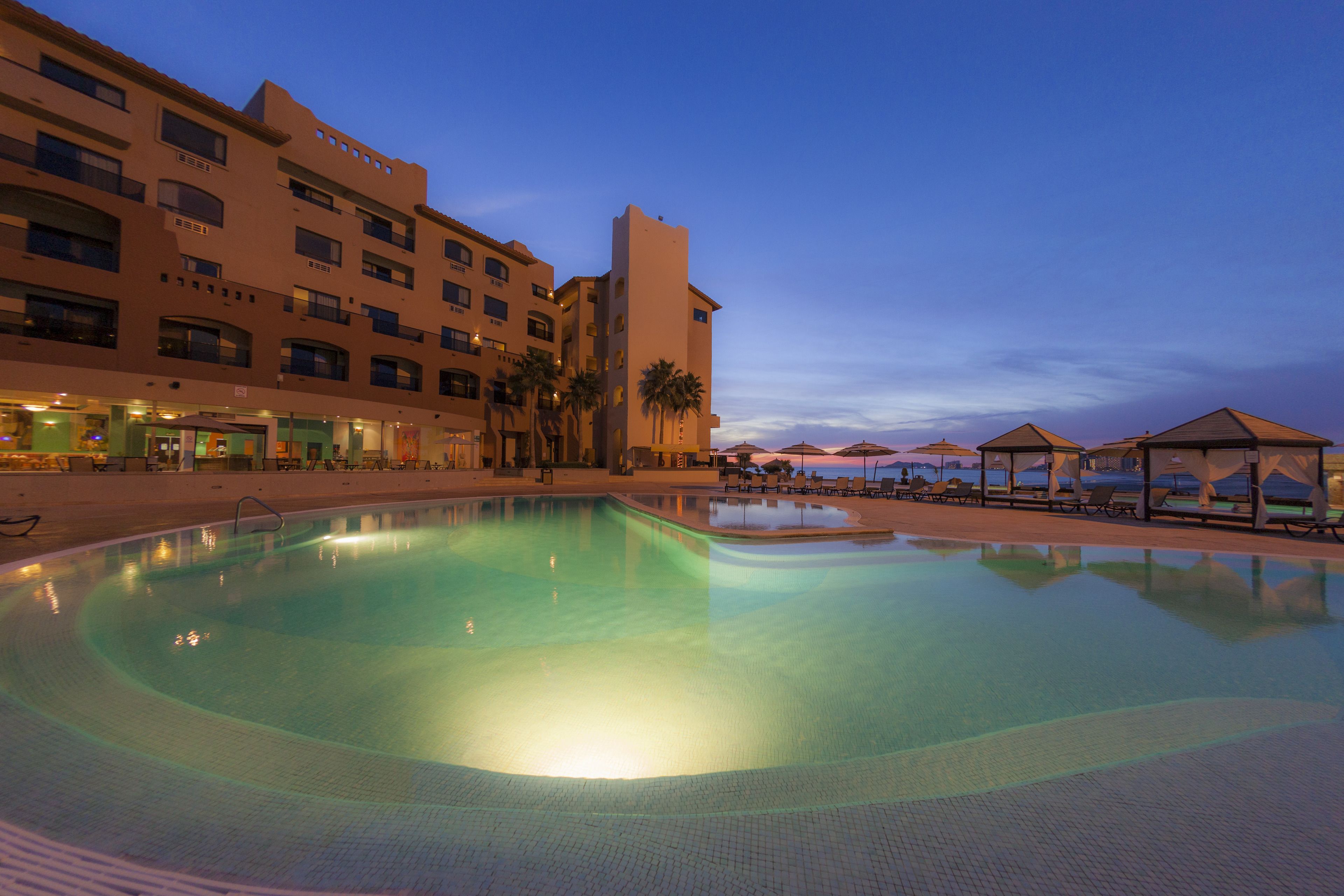 A swimming pool with a clear blue water surface, surrounded by a building with multiple balconies and palm trees.