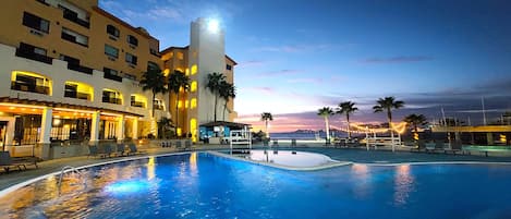 A hotel pool area with a large pool, lounge chairs, and palm trees.