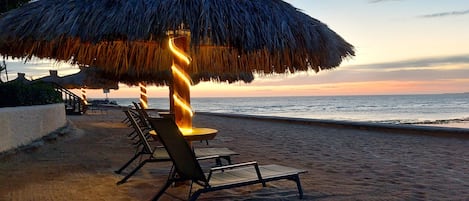 Eine Sitzgruppe am Strand mit einem Reedschirm und Liegestühlen, mit Blick auf das Meer.