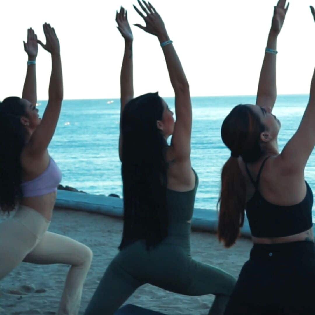 Three individuals performing a yoga pose on a beach with the ocean in the background.