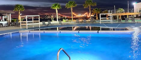 Una piscina con una escalera metálica, rodeada de palmeras y sillas de descanso.