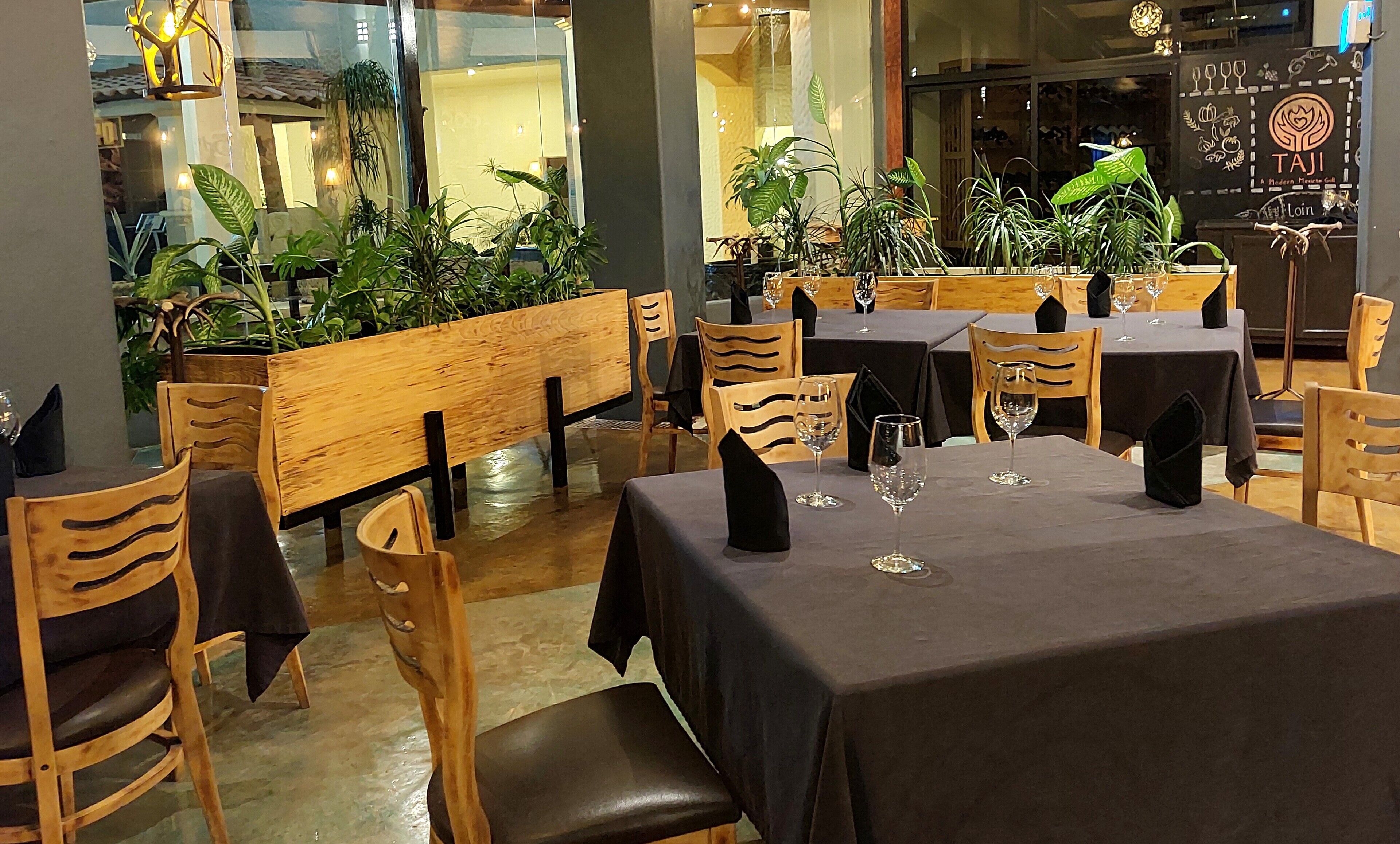 A restaurant interior with wooden tables and chairs, potted plants, and a black tablecloth.