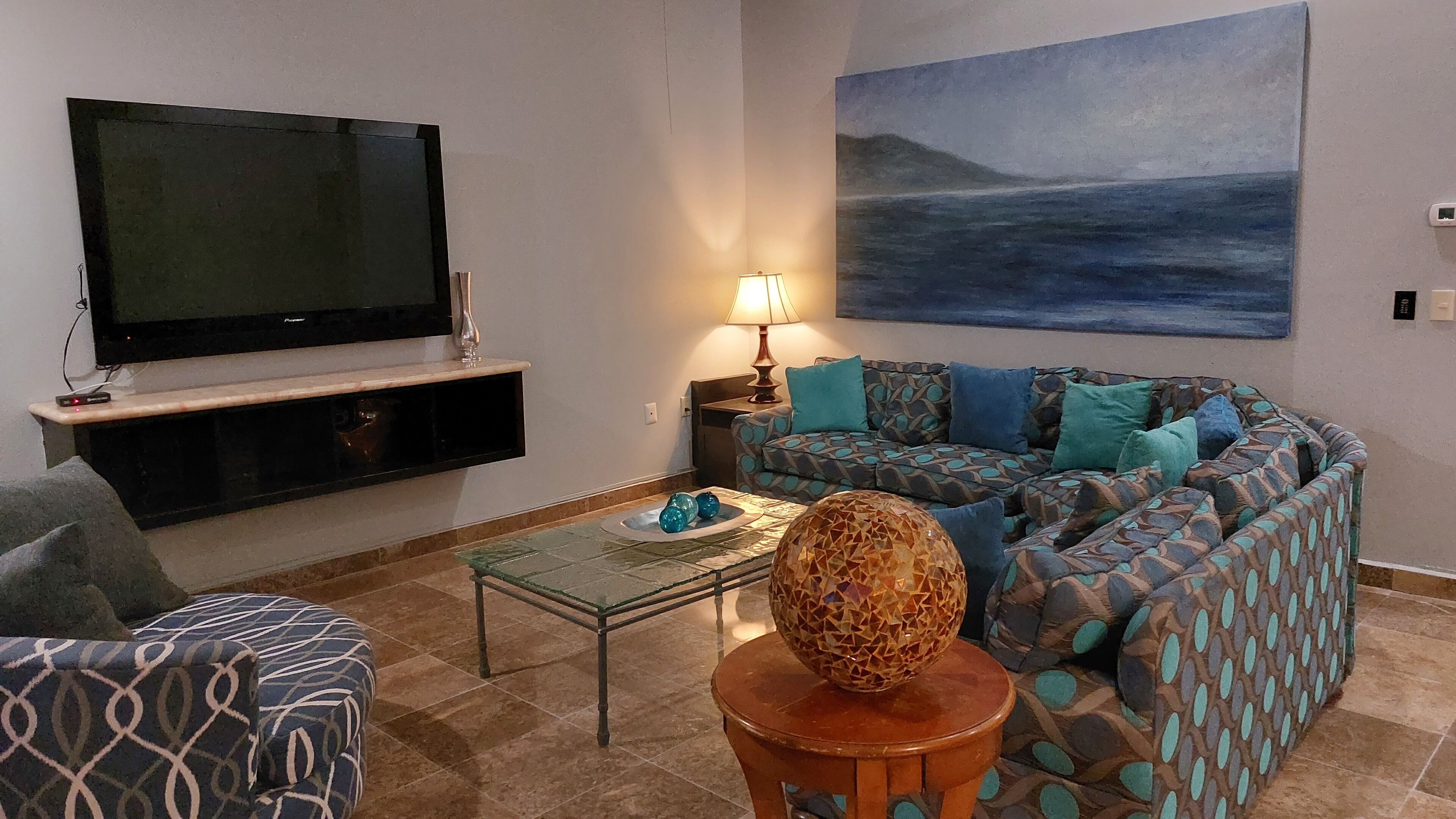 Uma sala de estar com televisão de tela plana, sofá estampado, mesa de centro com objetos decorativos e um quadro na parede.